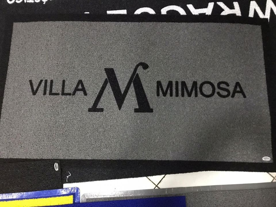 Capacho personalizado cinza com borda preta e a letra M estilizada ladeada pelo texto Villa Mimosa em preto, sobre piso de cerâmica, fotografia realista de ambiente residencial