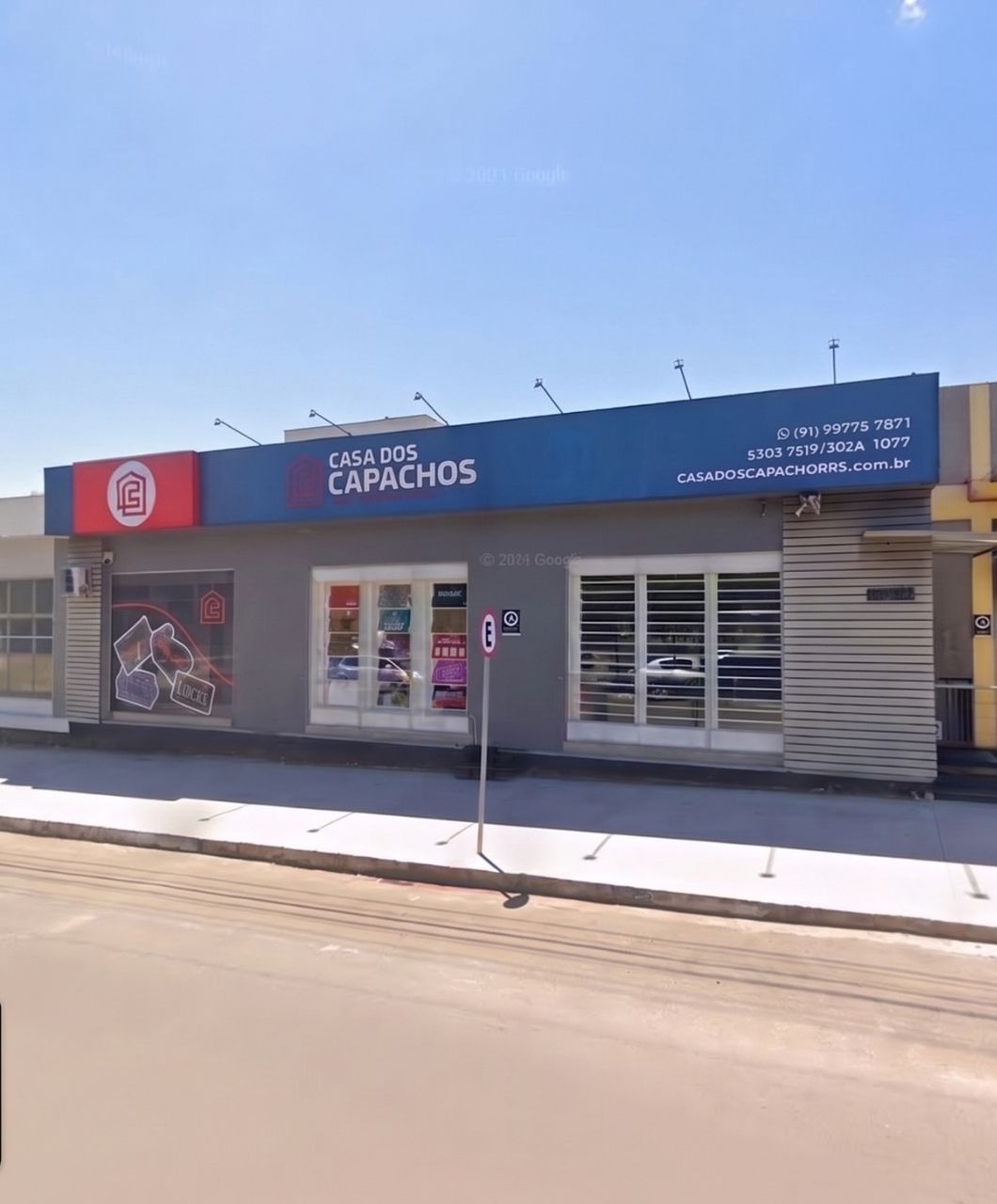 Fachada da loja Casa dos Capachos com letreiro azul e vermelho, entrada com vitrines de produtos, calçada em frente e céu azul claro, fotografia realista de rua comercial em dia ensolarado