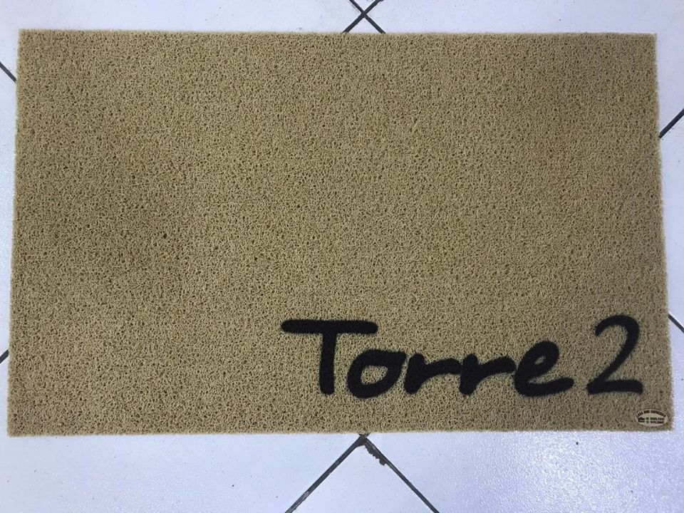 Capacho personalizado bege claro em fibra vinílica com o texto cursivo Torre 2 em preto no canto inferior direito, sobre piso de cerâmica clara, fotografia realista de ambiente residencial