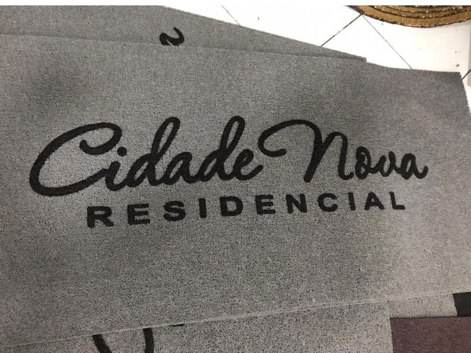 Capachos personalizados cinza empilhados com o texto cursivo Cidade Nova em preto e a palavra Residencial abaixo, sobre piso de cerâmica branca, fotografia realista de ambiente residencial de condomínio