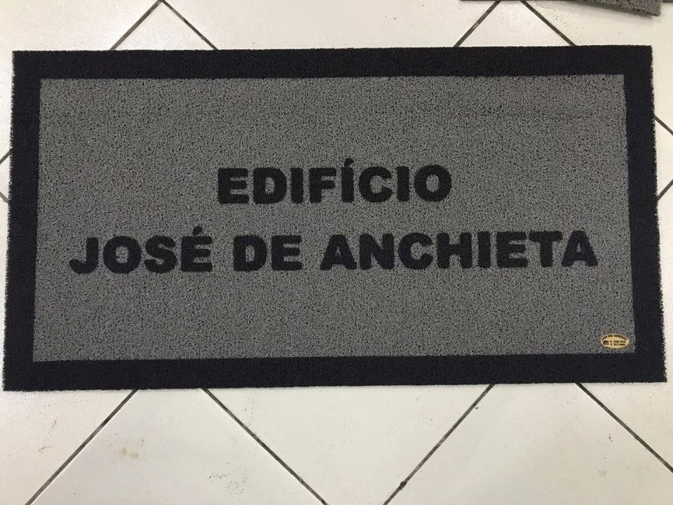 Capacho personalizado cinza com borda preta e o texto em duas linhas Edifício José de Anchieta em preto, sobre piso de cerâmica com juntas visíveis, fotografia realista de ambiente residencial