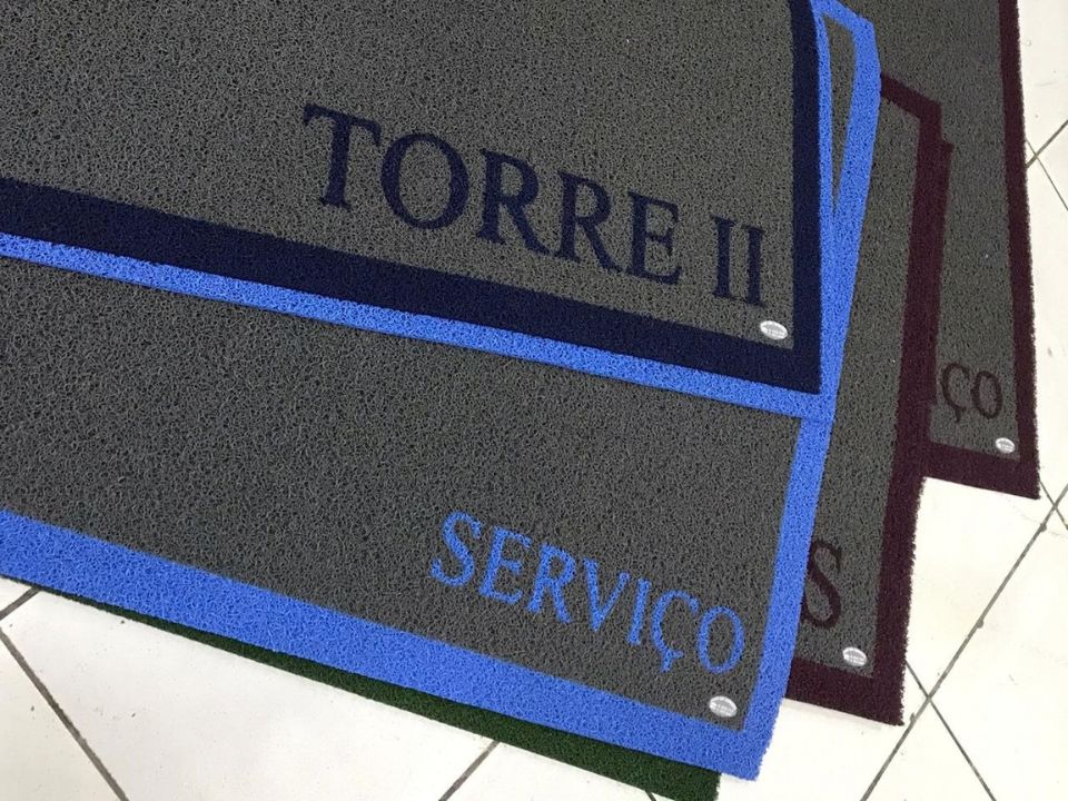 Capachos personalizados cinza com bordas coloridas em azul e vinho, com os dizeres Torre II e Serviço em destaque, dispostos lado a lado sobre piso de cerâmica clara, fotografia realista em ambiente de acesso de condomínio